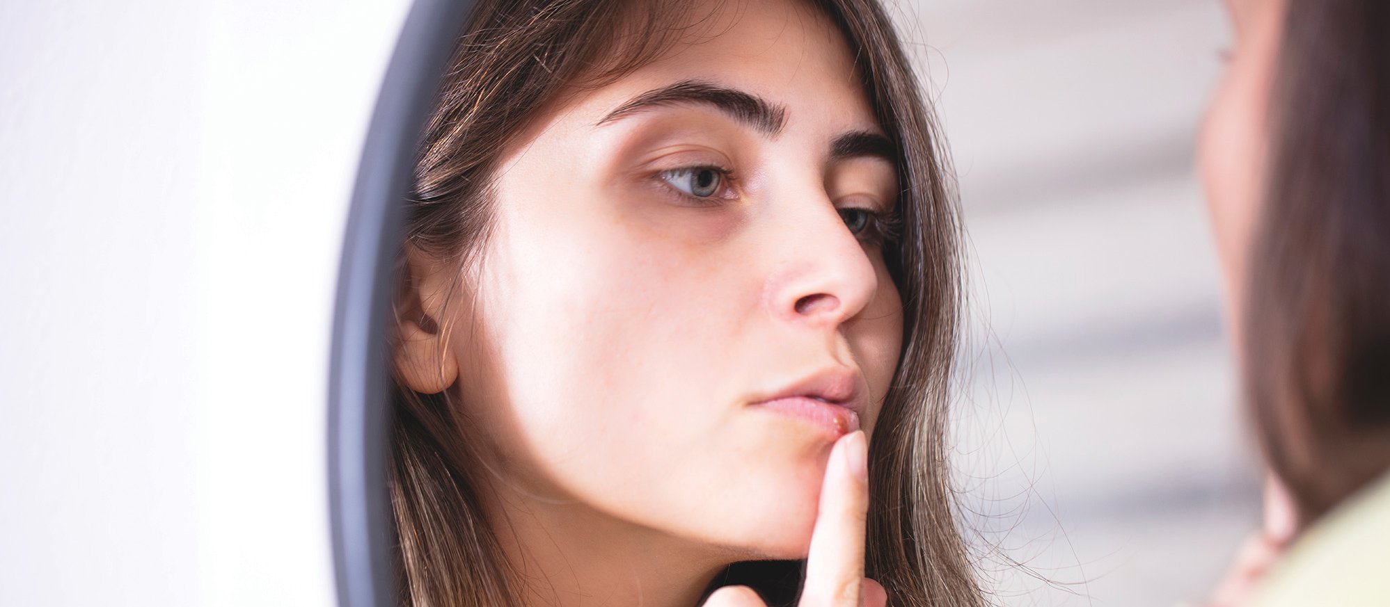 Woman looking at her cold sore in the mirror [GizemBR on iStock.com]