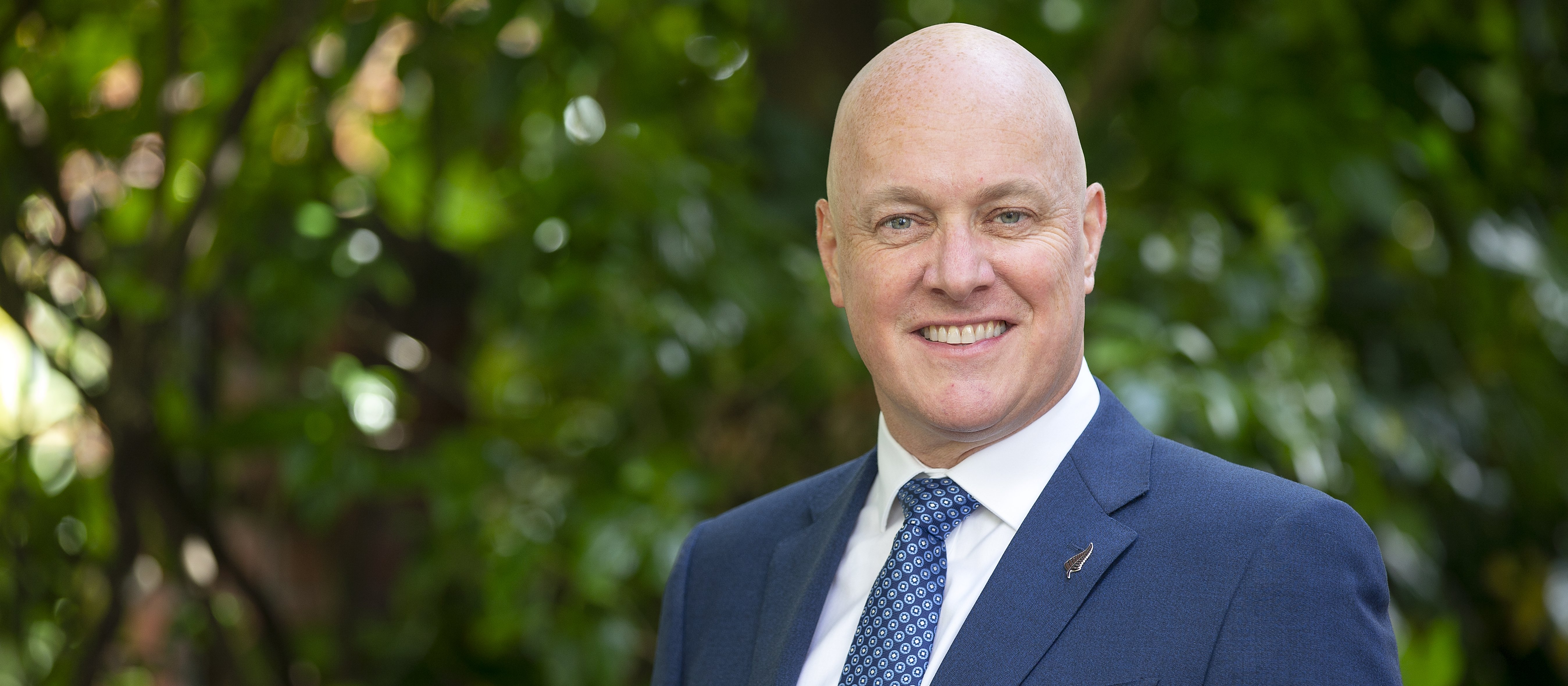 Christopher Luxon, in dark blue suit, white shirt and blue tie, trees in background