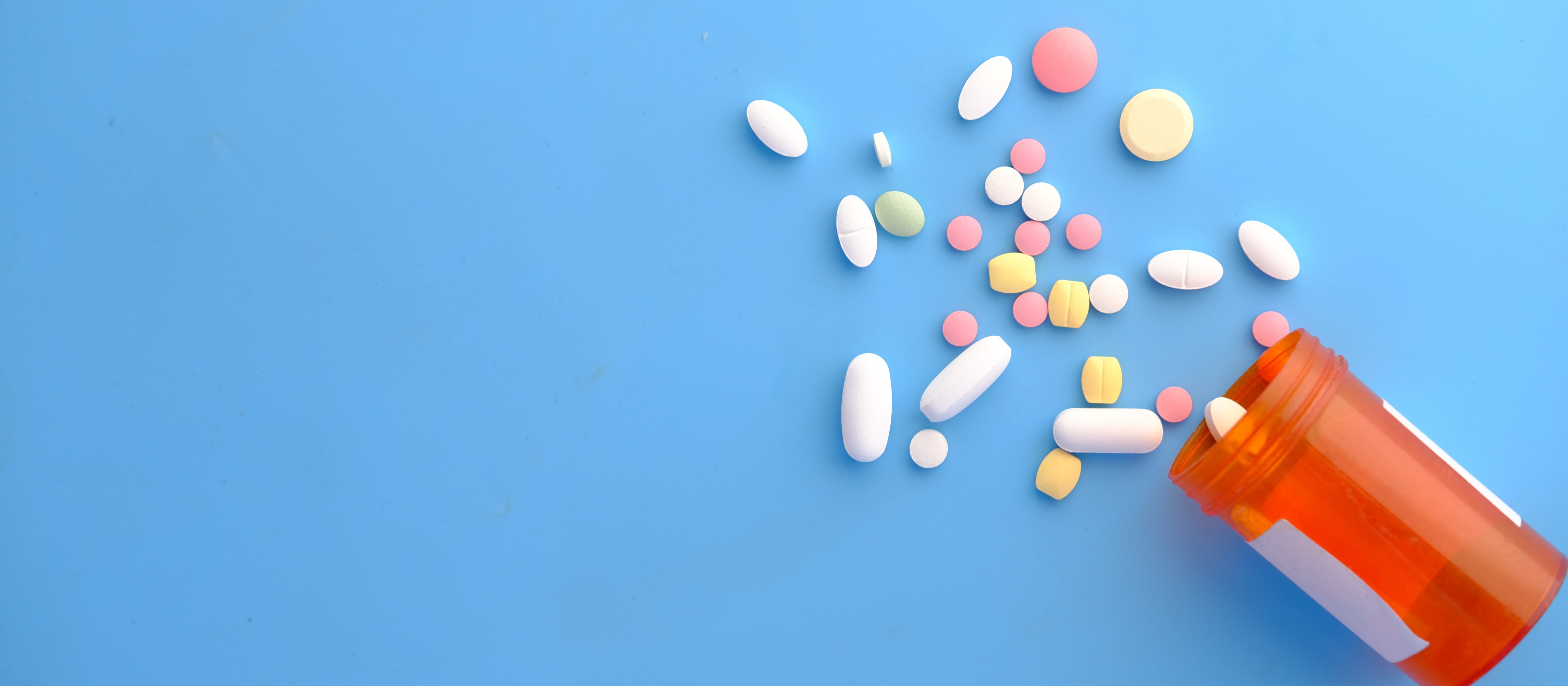 Different coloured pills spilling out of a medication jar on a blue background