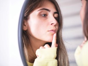 Woman looking at her cold sore in the mirror [GizemBR on iStock.com]