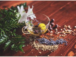 Image shows a clear glass cup of herbal tea, with flowers inside, on a wooden table strewn with herbs, leaves, and seeds