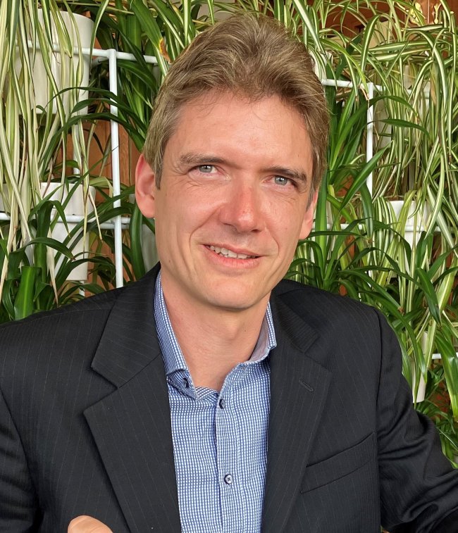 Image shows man in blue shirt and black pin-stripe blazer, with green leafy plants in background